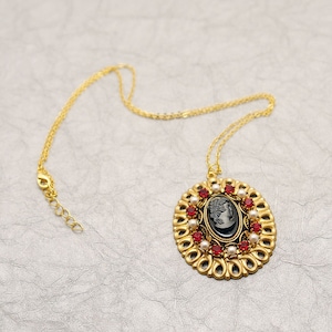 Könnte beinhalten: Eine goldene Kettenkette mit einem Kameenanhänger. Der Anhänger ist ein schwarzes Oval mit einem weißen Kamee eines Frauenprofils. Der Anhänger ist von einem goldenen Rahmen mit roten und weißen Strasssteinen umgeben.