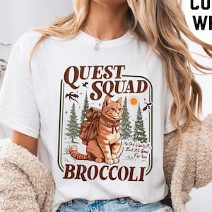 Op de afbeelding: Wit T-shirt met een afbeelding van een kat met een rugzak en de tekst "QUEST SQUAD BROCCOLI" in bruine letters. De tekst onder de kat luidt "Niemand wil het, maar het is goed voor je."