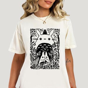 Puede incluir: Una camiseta de manga corta color crema con un estampado gr&aacute;fico negro de dos grandes criaturas forestales estilizadas rodeadas de &aacute;rboles, flores, mariposas y setas. El dise&ntilde;o tiene una est&eacute;tica de grabado en madera con l&iacute;neas marcadas.