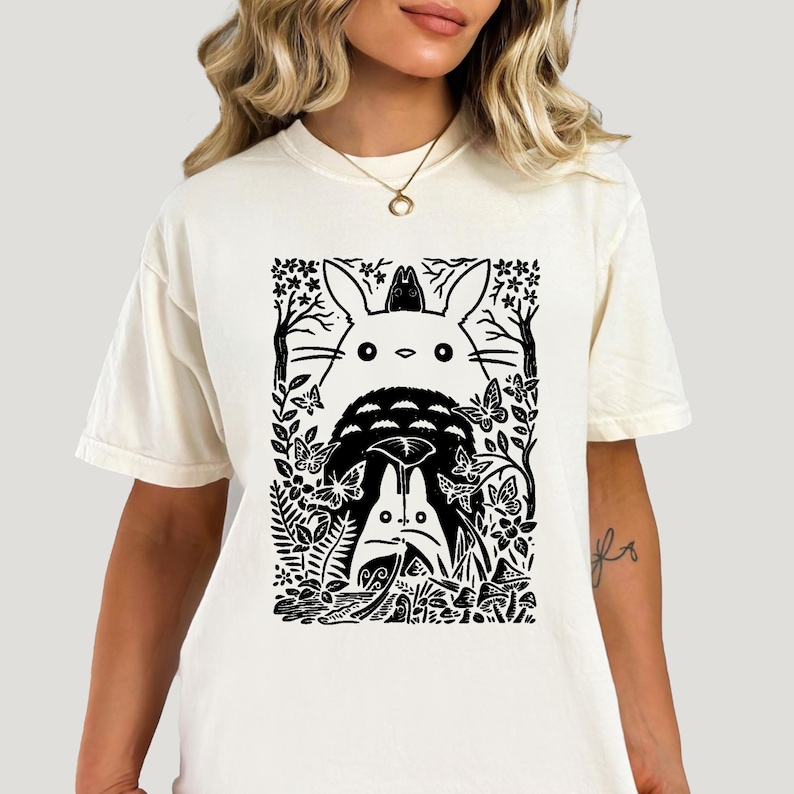 Puede incluir: Una camiseta de manga corta color crema con un estampado gr&aacute;fico negro de dos grandes criaturas forestales estilizadas rodeadas de &aacute;rboles, flores, mariposas y setas. El dise&ntilde;o tiene una est&eacute;tica de grabado en madera con l&iacute;neas marcadas.