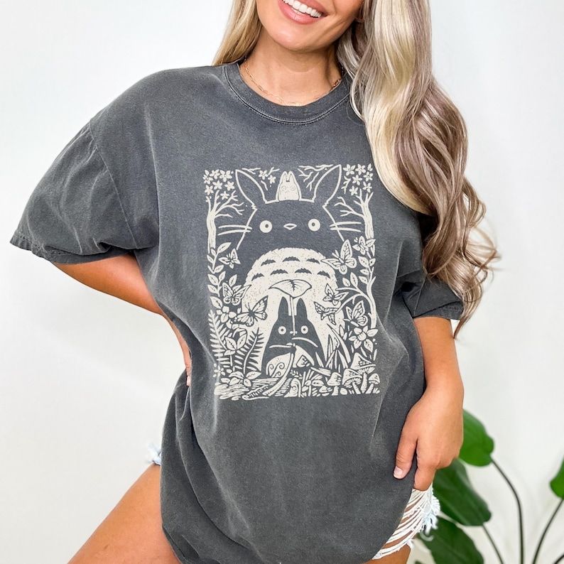 Puede incluir: Una camiseta gris carb&oacute;n de manga corta con un estampado gr&aacute;fico en color crema de dos criaturas del bosque rodeadas de mariposas, &aacute;rboles y follaje. El dise&ntilde;o incluye una criatura grande detr&aacute;s de una peque&ntilde;a que sostiene una hoja.