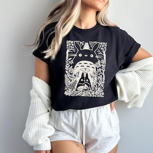 Puede incluir: Una camiseta negra de manga corta con un estampado gr&aacute;fico en blanco de un personaje de esp&iacute;ritu del bosque con orejas puntiagudas, un compa&ntilde;ero m&aacute;s peque&ntilde;o, mariposas y follaje. El dise&ntilde;o est&aacute; centrado en el pecho de la prenda.