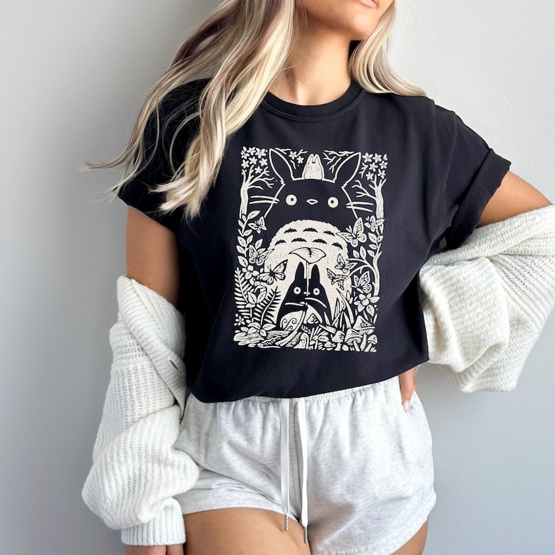 Puede incluir: Una camiseta negra de manga corta con un estampado gr&aacute;fico en blanco de un personaje de esp&iacute;ritu del bosque con orejas puntiagudas, un compa&ntilde;ero m&aacute;s peque&ntilde;o, mariposas y follaje. El dise&ntilde;o est&aacute; centrado en el pecho de la prenda.
