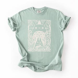 Puede incluir: Una camiseta de manga corta en color verde salvia claro con un estampado gr&aacute;fico color crema en la parte delantera. El dise&ntilde;o muestra una gran criatura del bosque con orejas puntiagudas, rodeada de esp&iacute;ritus, mariposas y follaje.