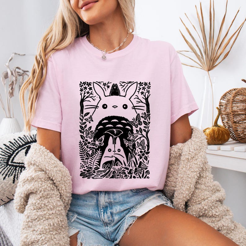 Puede incluir: Una camiseta de manga corta de color rosa claro con un estampado gr&aacute;fico negro de dos grandes criaturas parecidas a conejos rodeadas de mariposas, &aacute;rboles y follaje. El dise&ntilde;o incluye una peque&ntilde;a criatura posada sobre la cabeza del m&aacute;s grande.