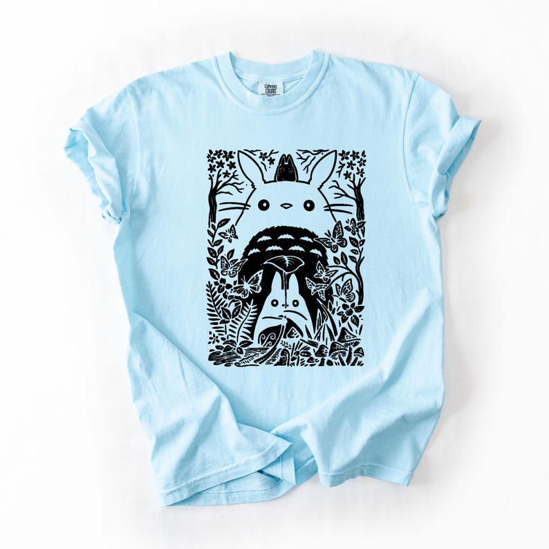 Puede incluir: Una camiseta azul claro con un estampado gr&aacute;fico negro de una criatura grande, redonda y parecida a un conejo con orejas largas, rodeada de criaturas del bosque m&aacute;s peque&ntilde;as, mariposas y follaje. El dise&ntilde;o est&aacute; enmarcado en un bloque rectangular.