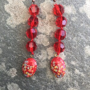 Könnte beinhalten: Ein Paar lange rote Ohrringe mit einem floralen Muster am unteren Ende. Die Ohrringe bestehen aus roten Glasperlen und haben einen Kupferhaken.
