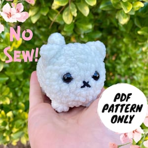以下が含まれることがあります： 黒い目をした猫の形をした白い毛糸のぬいぐるみ。おもちゃは手で持たれています。画像には「No Sew!」「PDF Pattern Only」の文字が見えます。