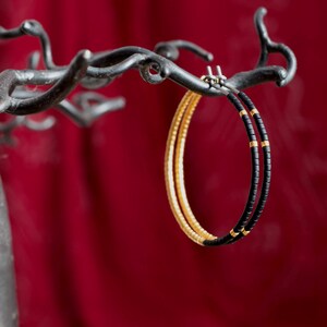 Könnte beinhalten: Ein Paar Creolen mit schwarzen und goldenen Perlen. Die Ohrringe hängen an einem schwarzen Metallständer.