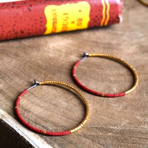 May include: A pair of gold hoop earrings with red beads. The earrings are made of small, round beads that are strung together to form a hoop shape. The beads are a mix of gold and red, and the earrings are a simple and elegant design.