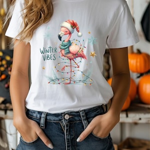 Peut inclure: T-shirt blanc avec un flamant rose portant un bonnet de Père Noël et une écharpe. Le flamant rose est entouré de lumières de Noël colorées et le texte "Winter Vibes" est imprimé sur le t-shirt.