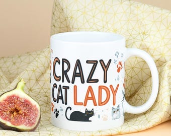 May include: White ceramic mug with the text "Crazy Cat Lady" in black and orange lettering. The mug features a black cat, a gray cat, and paw prints.