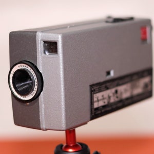 May include: A silver Kodak Ektamatic 35mm film camera with a black lens and a red button on the top. The camera is mounted on a red tripod.