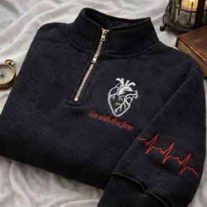May include: A dark grey sweatshirt with a quarter-zip neckline and a white embroidered anatomical heart design. The text "Go with the flow" is below the heart, and a red heartbeat line is on the sleeve.