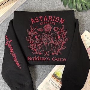 Puede incluir: Sudadera negra con un diseño bordado en rojo que representa a un personaje con un sol y una luna detrás. El texto "Astarion Approves" y "Baldur's Gate" también está bordado en rojo.