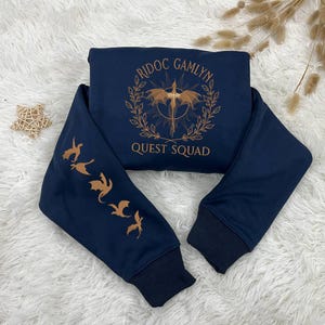 May include: A navy blue sweatshirt with a gold embroidered dragon and the text "Ridoc Gamlyn Quest Squad". The sleeves have gold embroidered dragons.