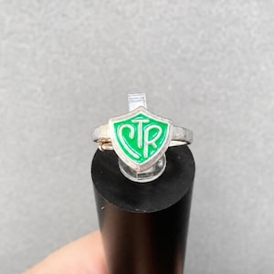 May include: A silver-toned metal ring featuring a shield-shaped emblem with a bright green enamel inlay. The center of the shield displays the letters CTR in a stylized, raised silver font. The band of the ring is plain and metallic in appearance.