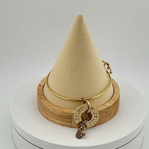 May include: A gold-colored bangle bracelet with a round charm and a mermaid pendant. The charm has engraved coordinates. The bracelet is displayed on a wooden stand with a beige cone-shaped top.
