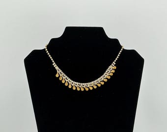 May include: A gold-toned necklace with clear and amber-colored rhinestones. The necklace is displayed on a black mannequin. The design features a series of clear rhinestones with amber teardrop-shaped accents.