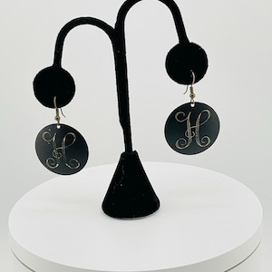 May include: A pair of black, circular earrings with a gold-colored letter "H" in a cursive font. The earrings hang from gold-colored hooks. The earrings are displayed on a black earring stand against a white background.