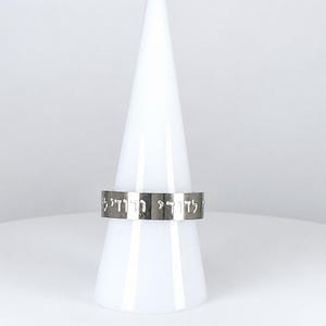May include: A silver-colored metal band ring featuring an engraved inscription in Hebrew characters. The ring has a smooth, polished finish and a flat profile, displaying the text clearly along the outer surface of the band.