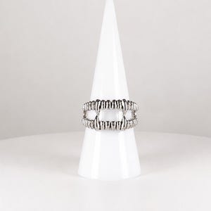 May include: A silver-toned metal ring featuring a design of two rows of teeth with prominent fangs. The band is constructed to resemble an open mouth, with the upper and lower teeth meeting at the sides and leaving a gap in the center.