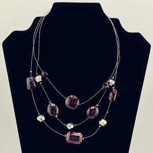 May include: A three-strand necklace featuring faceted, burgundy-colored beads in various shapes, including rectangles, ovals, and circles. Silver-toned, geometric accent beads are interspersed throughout the design. The necklace is displayed on a black mannequin.