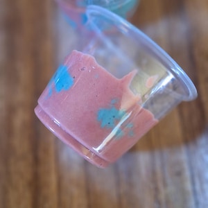 May include: Two clear plastic cups with lids. One cup contains a pink smoothie with blue swirls. The other cup contains a blue smoothie with pink swirls.