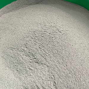 May include: A close-up shot of a pile of fine, light gray powder. The powder has a textured, granular appearance, filling the frame. The background is a bright green, creating a contrast with the gray powder.