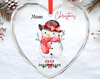 May include: A heart-shaped clear glass ornament featuring a watercolor snowman wearing a brown hat with a red bow and a red polka-dot scarf. The text reads Merry Christmas and 2025, accented by a small red bow and green holly leaves.