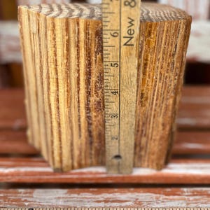 Puede incluir: Un bloque de madera natural con veta visible, de aproximadamente 15 cm de altura, se muestra con una regla de madera. La regla, marcada en pulgadas, está colocada junto al bloque. El bloque está sobre una superficie roja.