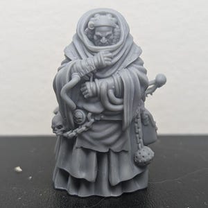 Op de afbeelding: Een grijs 3D-geprint miniatuur van een gekapte figuur met een schedel en een ketting die aan zijn riem is bevestigd. De figuur draagt een lange robe en houdt een staf in zijn hand.