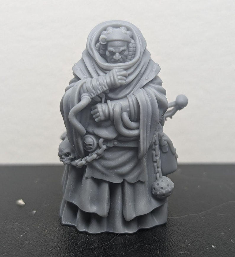 Op de afbeelding: Een grijs 3D-geprint miniatuur van een gekapte figuur met een schedel en een ketting die aan zijn riem is bevestigd. De figuur draagt een lange robe en houdt een staf in zijn hand.