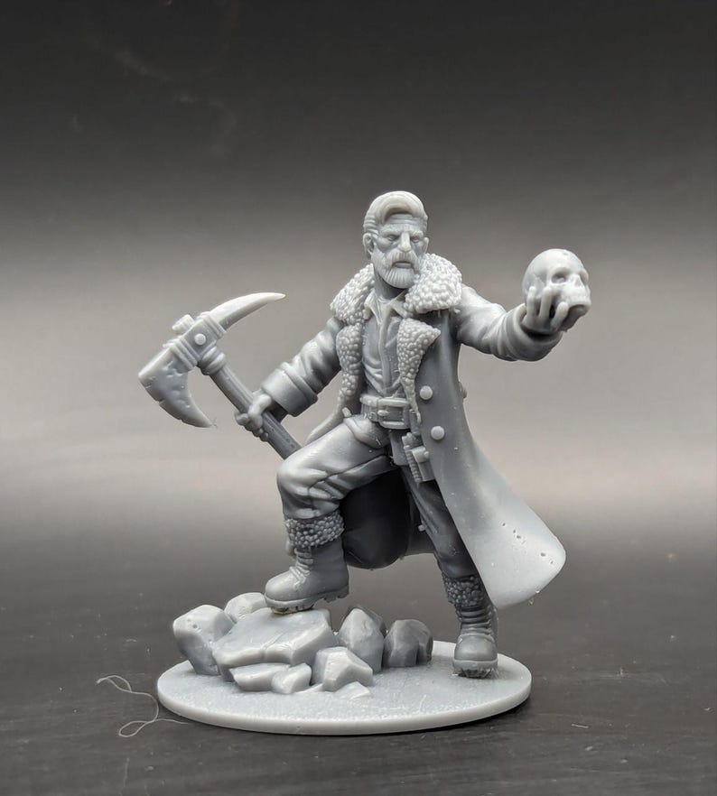 Op de afbeelding: Een grijs 3D-geprint miniatuur van een persoon die een lange jas draagt, een schedel in de ene hand en een houweel in de andere hand vasthoudt. Het miniatuur staat op een rotsachtige basis.