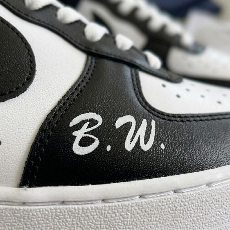 May include: Close-up of a black and white sneaker. The shoe features a black swoosh, black leather accents, and the initials "B.W." in white script. The laces and sole are white.