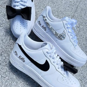 Op de afbeelding: Een paar witte sneakers met zwarte swooshes. E&eacute;n sneaker heeft een zwarte strik en de tekst "Mr. Kelly" op de zijkant. De andere sneaker heeft een wit satijnen lint en de tekst "Mrs. Kelly" op de zijkant. De sneaker heeft ook een glinsterende, met strass bezette swoosh.