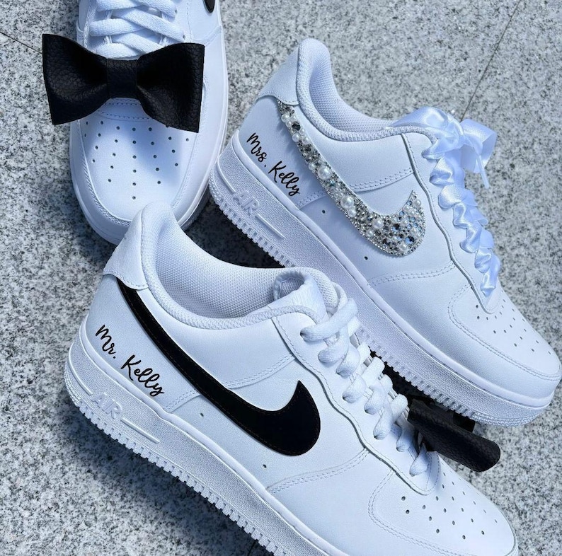 Op de afbeelding: Een paar witte sneakers met zwarte swooshes. E&eacute;n sneaker heeft een zwarte strik en de tekst "Mr. Kelly" op de zijkant. De andere sneaker heeft een wit satijnen lint en de tekst "Mrs. Kelly" op de zijkant. De sneaker heeft ook een glinsterende, met strass bezette swoosh.