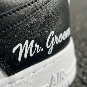 May include: Close-up of a black and white sneaker. The words "Mr. Groom" are written in white script on the black leather heel. The white sole has the word "AIR" printed on it.