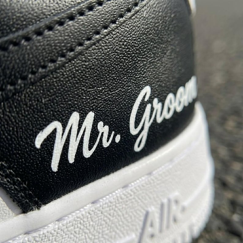 May include: Close-up of a black and white sneaker. The words "Mr. Groom" are written in white script on the black leather heel. The white sole has the word "AIR" printed on it.