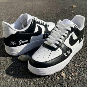 May include: A pair of white and black sneakers with the words "Mr. Groom" and initials on the sides. The sneakers have white laces and soles. The shoes are sitting on a dark gray surface.