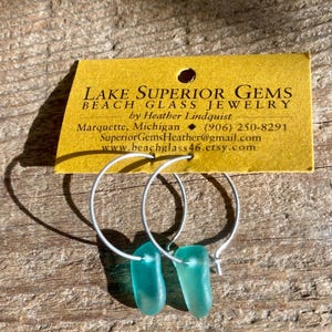 May include: A pair of silver hoop earrings with sea glass pendants. The sea glass is a translucent teal color. The earrings are displayed on a yellow tag with the text "LAKE SUPERIOR GEMS BEACH GLASS JEWELRY".