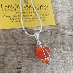 May include: A silver necklace with a pendant made of orange sea glass. The sea glass is wrapped in silver wire. The necklace is on a wooden surface. A yellow tag with text is in the background.