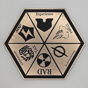 May include: A hexagonal metal game piece with black and gold accents. The piece has six sides, each with a different symbol and text. The symbols include a lightning bolt, a shield, a cloud with lightning bolts, a circle with a line through it, a radiation symbol, and a wreath with a cross. The text on the sides includes "Energy", "Experience", "Storm", "Poison Counter", "Raid", and "Tax/Commander".