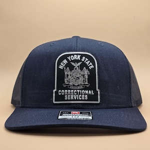 May include: A navy blue trucker hat with a black mesh back. The hat features a white patch with the text "New York State Correctional Services" and the New York State seal.