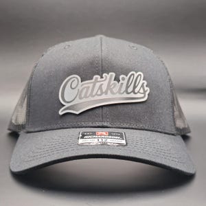 May include: A charcoal gray trucker hat with a mesh back and a curved brim. The hat features the word "Catskills" in a silver, stylized font on the front. A small tag with text is visible on the brim.