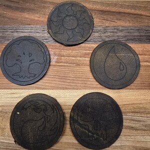 May include: Five round leather coasters with different symbols. The symbols are a sun, a tree, a drop of water, a wave, and a skull.