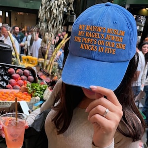 Op de afbeelding: Een blauwe honkbalpet met oranje geborduurde tekst: MY MAYOR'S MUSLIM, MY BAGEL'S JEWISH, THE POPE'S ON OUR SIDE, KNICKS IN FIVE. De pet heeft een gestructureerde kroon, een gebogen klep en een knoop aan de bovenkant, gemaakt van katoenen stof.