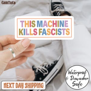 Op de afbeelding: Een rechthoekige sticker met de tekst "THIS MACHINE KILLS FASCISTS" in kleurrijke blokletters. De sticker wordt vastgehouden door een hand, met een ring om de vinger. De achtergrond omvat een paar zwarte sneakers en een wit oppervlak.