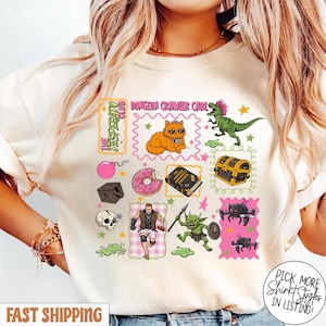 Peut inclure: T-shirt beige avec des graphismes de dessins animés colorés. Le motif comprend un chat avec des lunettes de soleil, un dinosaure, un coffre au trésor et le texte "Dungeon Crawler Carl". D'autres graphismes incluent un crâne, un beignet et un gobelin. Le t-shirt a le texte "The Desperado Club".