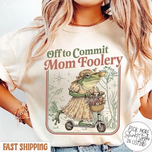 May include: A cream-coloured t-shirt with the text "Off to Commit Mom Foolery." The graphic shows a frog in a dress and hat riding a scooter, with a basket of flowers. The design is framed in a retro style.